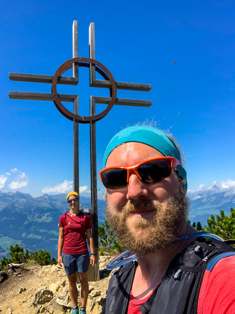 Wanderungen in Liechtenstein. Selfie von Melanie und Julian mit Gipfelkreuz der Alpsitz. Der Himmel ist kräftig blau.