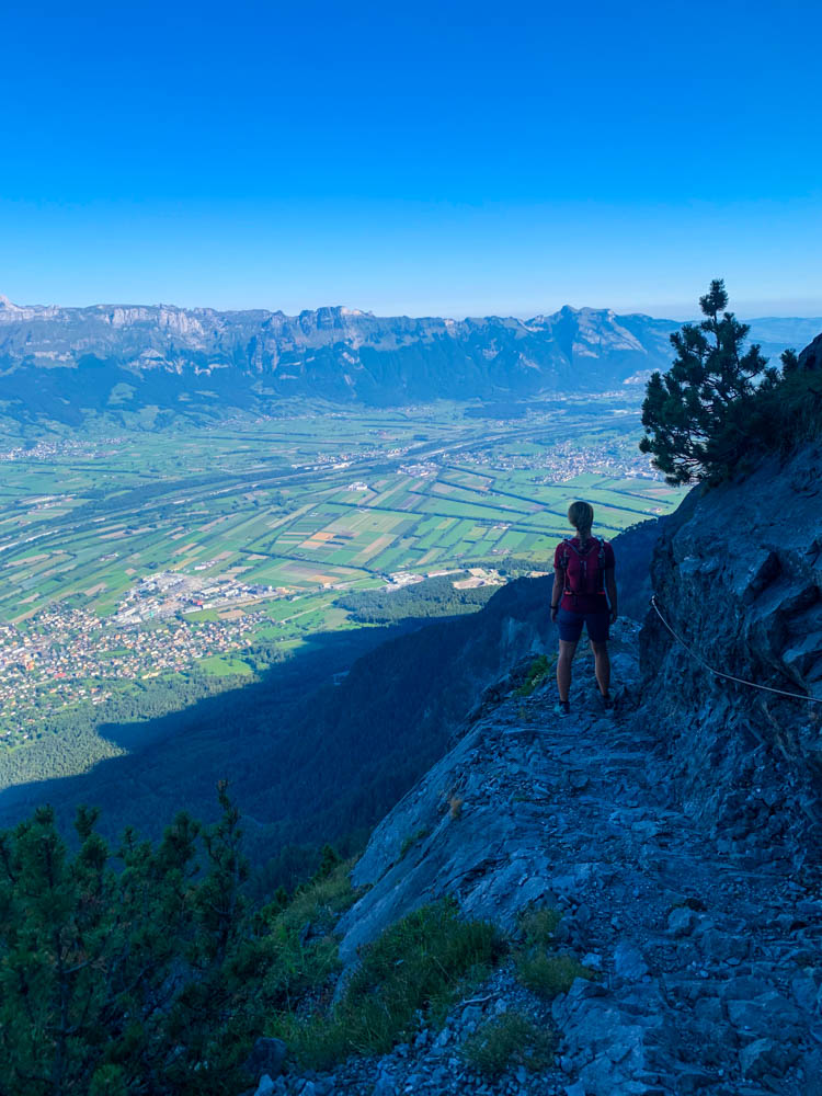 Wanderungen in Liechtenstein. Melanie im Aufstieg auf dem Fürstensteig im felsigen Gelände auf einem schmalen Pfad. Der Himmel ist kräftig blau. Melanie blickt auf die Felder im Tal sowie die gegenüberliegenden Berge.