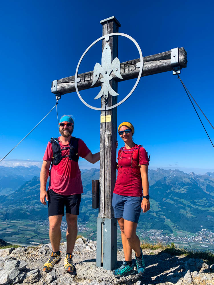Wanderungen in Liechtenstein. Melanie und Julian am Kuegrat. Das Gipfelkreuz steht zwischen den beiden, der Himmel ist kräftig blau.