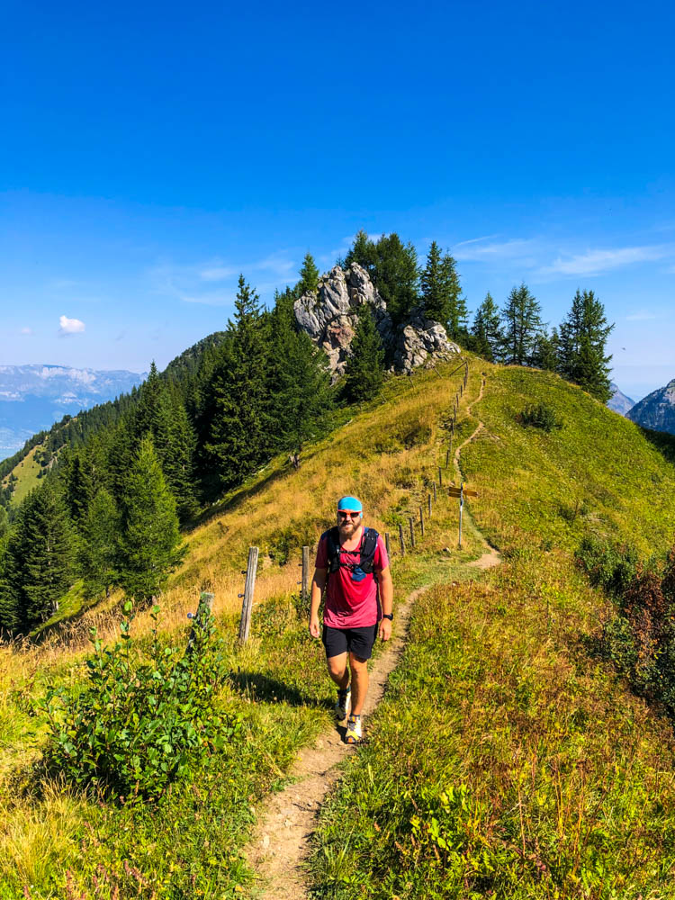 Wanderungen in Liechtenstein. Julian beim wandern auf einem schmalen Bergpfad entlang eines Zaunes. Der Himmel ist kräftig blau.