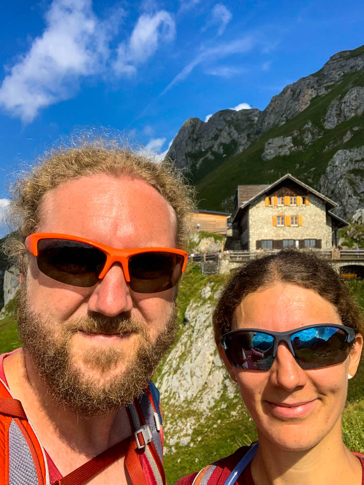 Selfie von Melanie und Julian im Tannheimer Tal. Hinter ihnen ist die Bad Kissinger Hütte zu sehen.