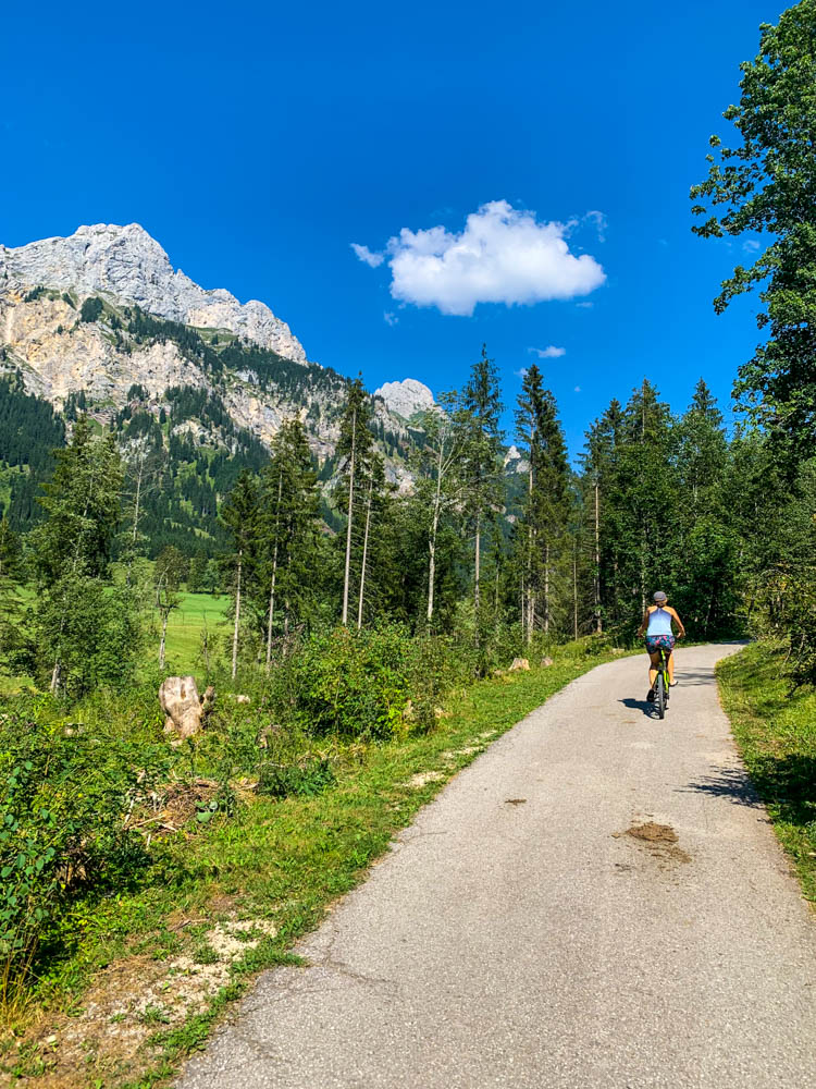 Melanie fährt mit dem Fahrrad auf einem Forstweg durch ein Waldabschnitt. Links sind Berg zu sehen.