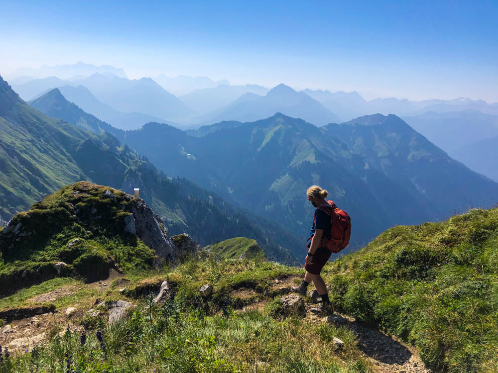 Touren im Tannheimer Tal - Julian im Abstieg im Gipfelbereich der Rote Flüh. Im Hintergrund ist ein Bergpanorama zu sehen, der Himmel ist kräftig blau.