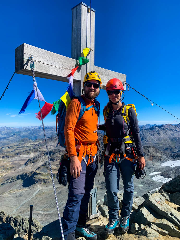 Gipfelfoto mit Gipfelkreuz von Melanie und Julian am Großen Piz Buin. Sie strahlen in die Kamera. Hinter ihnen sind einige Berge zu sehen sowie der kräftig blaue Himmel