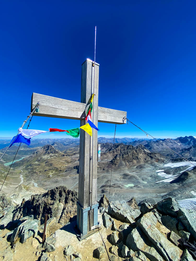 Gipfelkreuz am Großen Piz Buin. Es weht eine bunte Gebetsfahne im Wind, es sind einige Berge im Tal zu sehen sowie der kräftig blaue Himmel.
