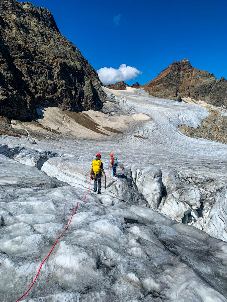 Melanie wandert über eine der vielen Spalten im Ochsentaler Gletscher nach der Piz Buin Besteigung. Es sind einige Spalten auf dem Bild zu sehen, ein paar Berge sowie der kräftig blaue Himmel