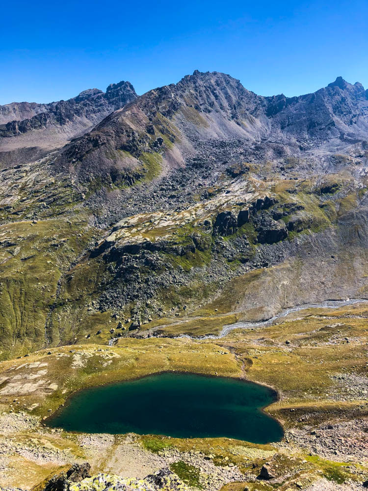 Wanderung Vorarlberg Radschulter und Radsattel. Im Tal ist ein See zu sehen, der die Form eines Herzes hat. Berge sowie der kräftig blaue Himmel vervollständigen das Bild.