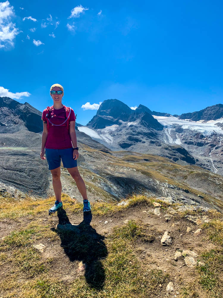 Melanie grinst in die Kamera und posiert mit der Berglandschaft im Hintergrund um die Wette. Piz Buin und Gletscher, sowie der kräftig blaue Himmel sind zu sehen.