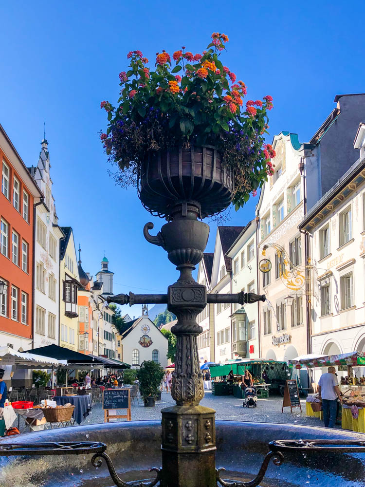 Innenstadt Feldkirch. Im Zentrum des Bildes ist ein Brunnen zu sehen, dahinter ein Markt mit kleinen Ständen, die Häuserfassaden sowie der strahlend blaue Himmel.