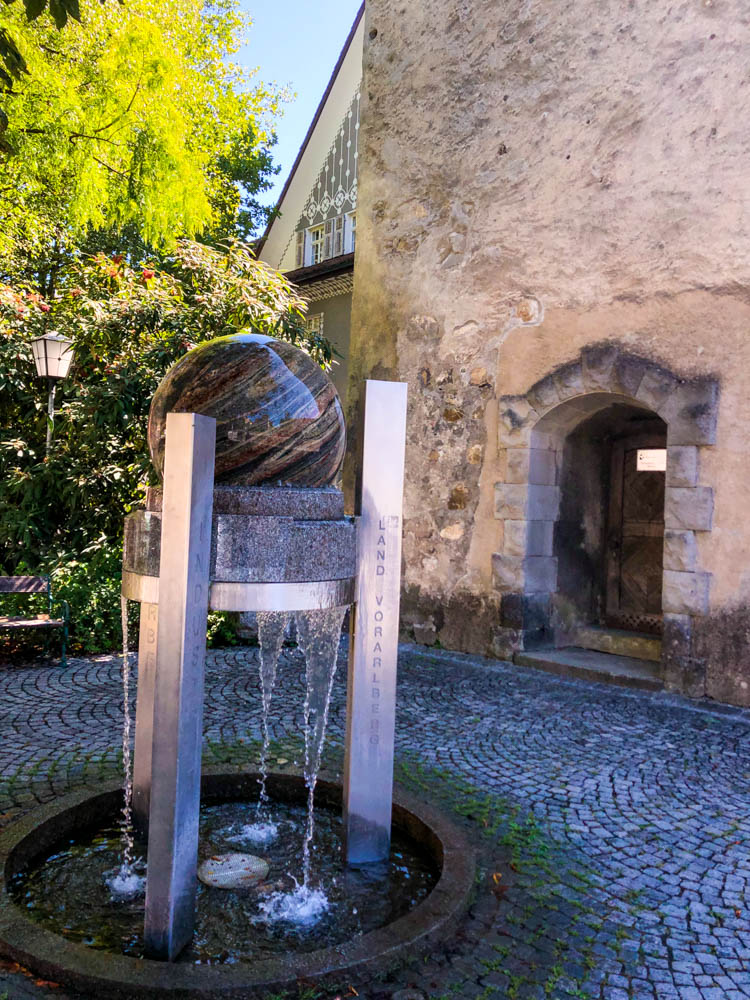 Es steht eine Kugel auf einem Ständer aus Stein in Feldkirch und es läuft Wasser unten raus. Es ist die Gravur Land Vorarlberg zu sehen.