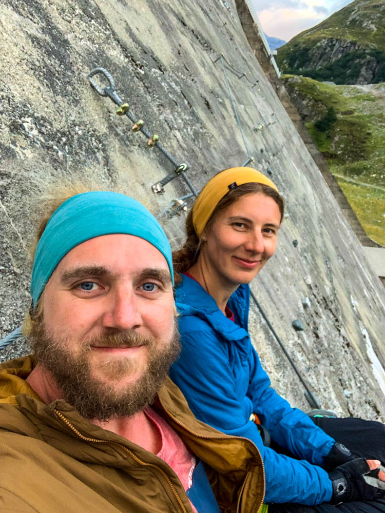 Selfie von Melanie und Julian im Silvretta Stausee Klettersteig. Sie sitzen gerade auf einer Bank, welche im Klettersteig befestigt ist. Hinter ihnen ist die Staumauer mit Drahtseilen und Tritten zu sehen.
