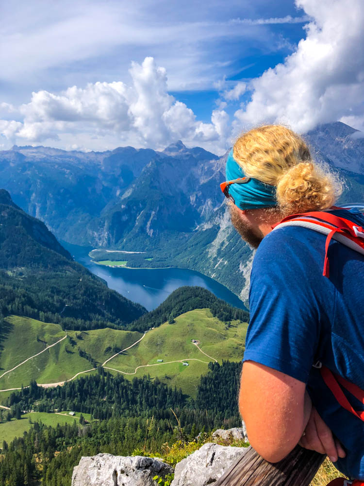 Julian blickt von einer Aussichtsplattform am Jenner hinab auf den Königssee sowie die Berchtesgadener Alpen. Watzmann und eine schöne Wald- und Wiesenlandschaft sind zu sehen. Der Himmel ist leicht bewölkt.