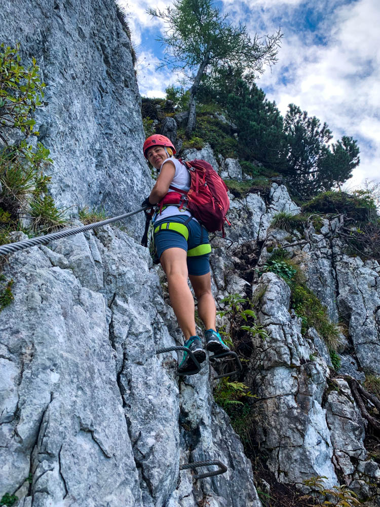 Melanie im Schützensteig Klettersteig in den Berchtesgadener Alpen. Sie steht auf Eisenstiften und grinst in die Kamera. Felsiges Gelände mit Drahtseil abgesichert.