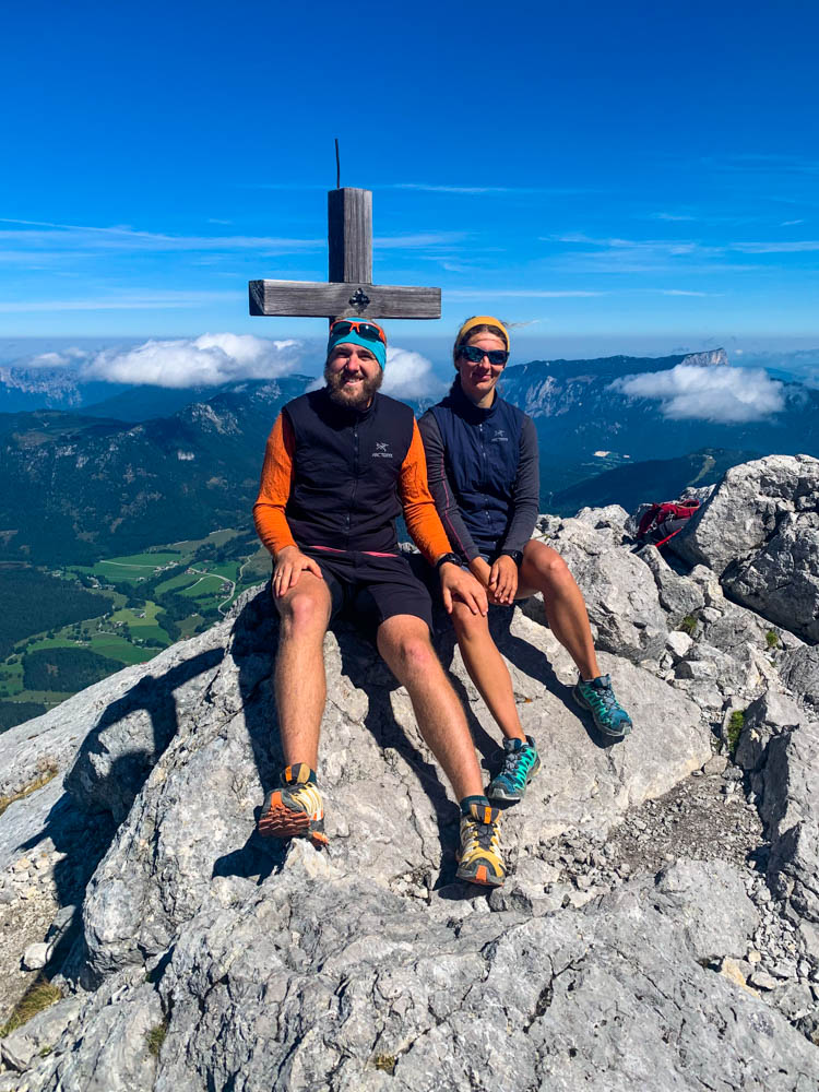 Gipfelfoto mit Gipfelkreuz von Melanie und Julian. Das Gipfelkreuz der Schärtenspitze ist ziemlich klein, deshalb sitzen die beiden daneben, um es nicht zu überragen. Blick ins Tal sowie auf die Berglandschaft der Berchtesgadener Alpen, der Himmel ist kräftig blau.