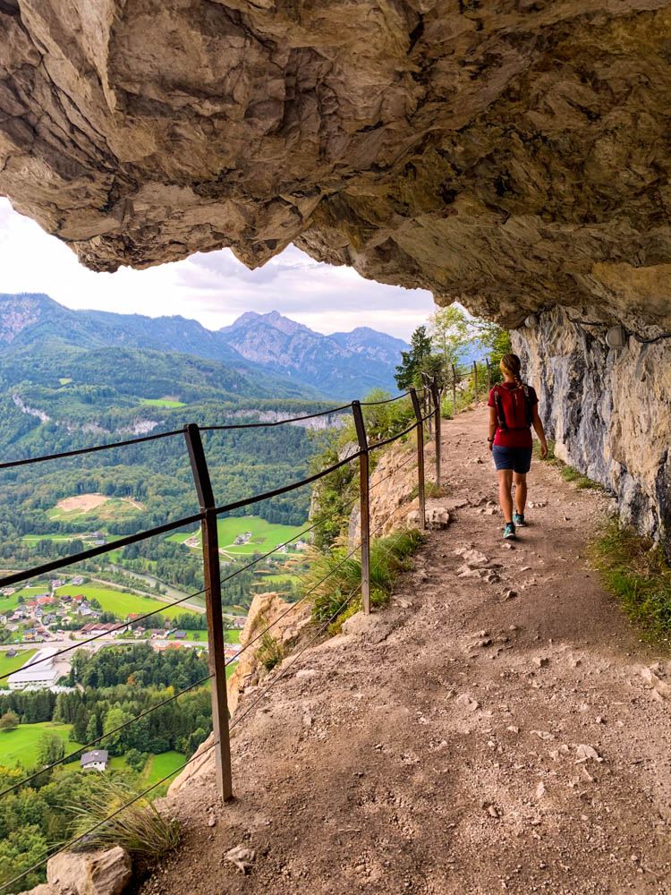 Es ist ein Pfad in der Ewigen Wand zu sehen, welcher vom Fels überragt wird. Melanie wandert unter dem Felsvorsprung, links vom Bild Blick ins Tal und auf die Berge. Querfeldein durch Österreich