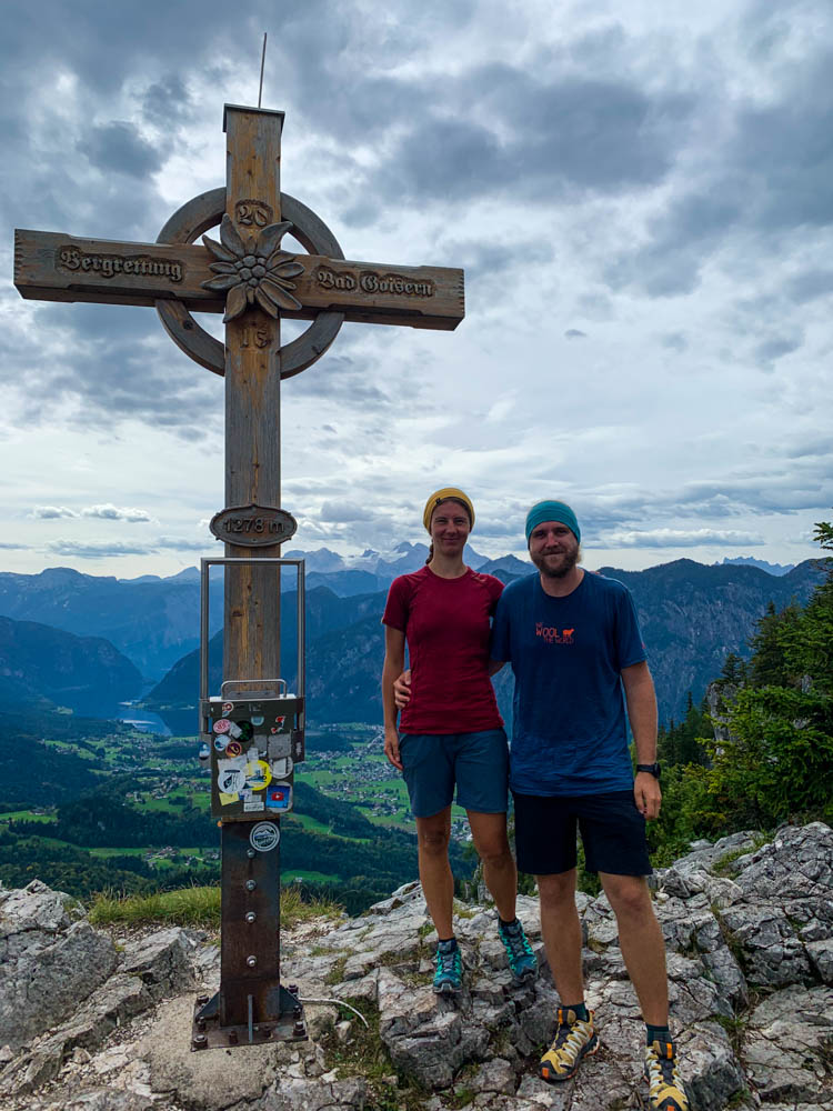 Aufnahme von Melanie und Julian mit Gipfelkreuz am Predigstuhl. Im Hintergrund ist der Dachstein zu sehen. Querfeldein durch Österreich