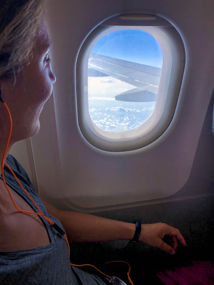 Melanie blickt aus dem Fenster eines Flugzeugs. Sie sieht einen Flügel des Flugzeugs, das Wolkenmeer sowie den blauen Himmel