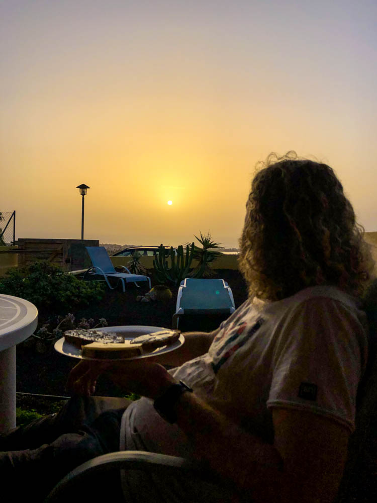 Julian sitzt auf einer Terrasse, die Sonne geht gerade auf und er blickt auf das Meer am Horizont. Calima auf Fuerteventura: der Himmel ist orange-braun verfärbt und die Sicht ist eingeschränkt