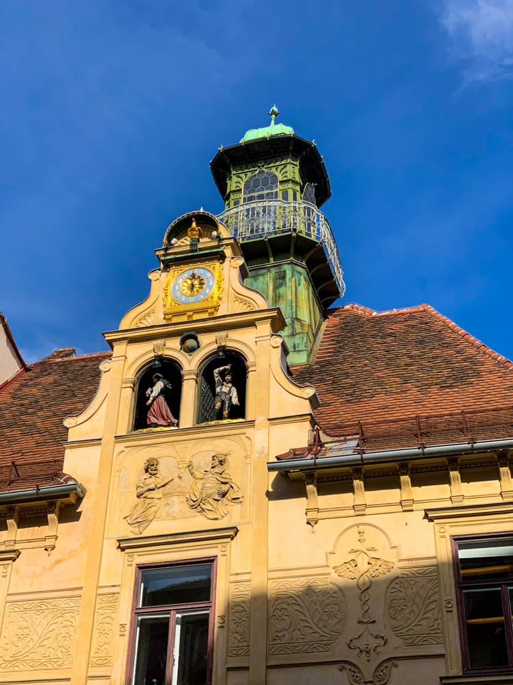 Glockenspielplatz mit steirischem Tanzpaar (Figuren), welche in Giebelfenstern tanzen. Der Himmel ist kräftig blau.