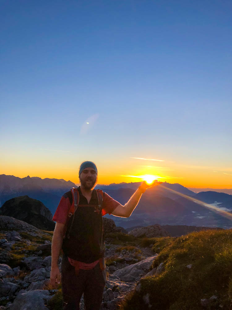 Julian beim Sonnenaufgang in den Berchtesgadener Alpen. Er hält die Hand auf und es sieht aus als ob er die Sonne in der Hand hätte. Orange-rote Töne über den Gipfeln.
