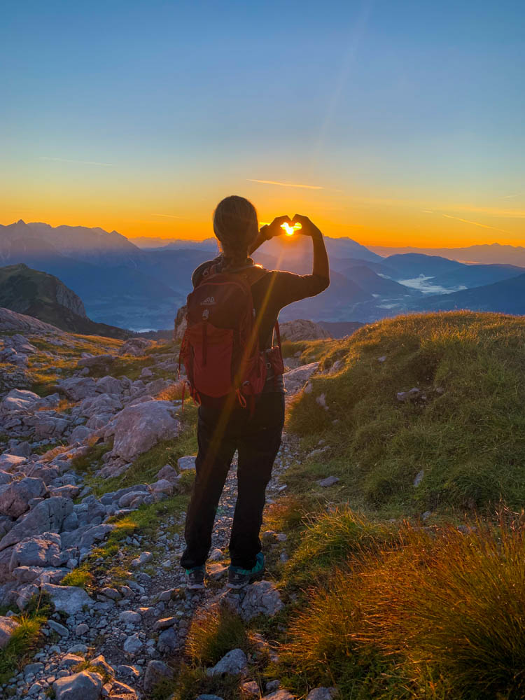 Melanie formt mit ihren Händen ein Herz und schließt darin die Sonne ein. Sonnenaufgang in den Berchtesgadener Alpen, über den Bergen ist alles orange-rot verfärbt. Der Himmel ist wolkenfrei. Wanderung Hochkönig