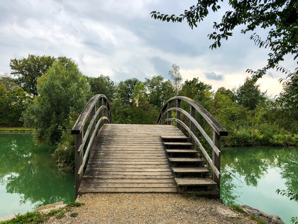 Badesee in Österreich. Es führt eine Brücke über den See, welcher von Bäumen umgeben ist. Das Wasser schimmert türkisfarben. Querfeldein durch Österreich
