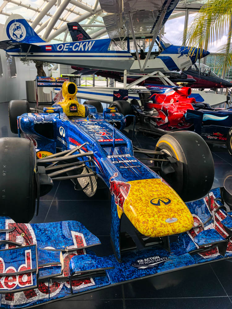 Formel-1 Wagen mit Red Bull Werbung im Hangar 7 in Salzburg ausgestellt. Dahinter sind ein Sportflugzeug sowie ein weiterer Formel-1 Wagen zu sehen.