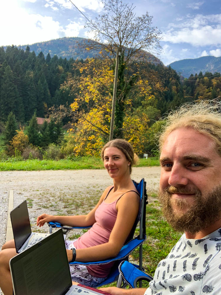 Melanie und Julian sitzen vor Van Vivaldi und haben einen Laptop auf dem Schoß. Sie grinsen beide in die Kamera. Im Hintergrund ein Wald Sloweniens