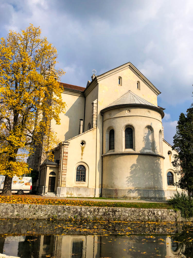 Kirche in Slowenien, welche sich in einem Fluss spiegelt. Der Himmel ist bewölkt