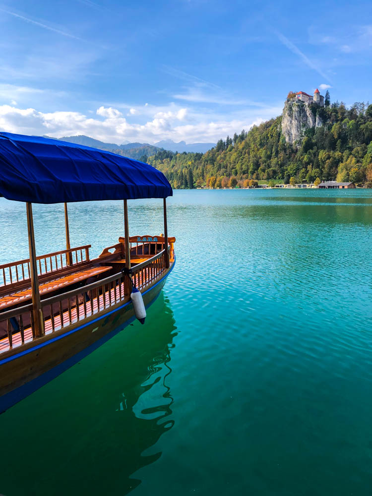 Holzboot mit blauem Dach liegt im Bleder See, welcher türkisfarben schimmert. Gegenüber des Sees ist die Burg von Bled zu sehen. Roadtrip Slowenien bei blauem Himmel