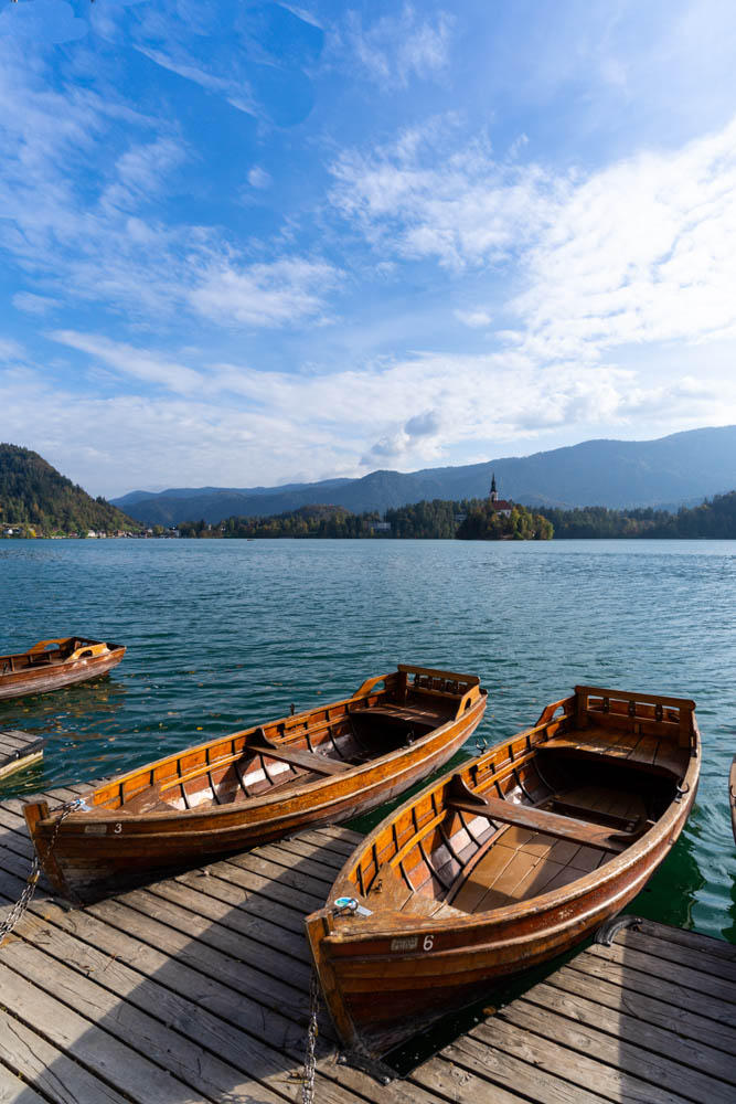Mehrere Holzboote liegen am Ufer des Bleder Sees. Die Kirche auf der Insel des Sees ist zu sehen, sowie eine paar Schleierwolken am Himmel. Roadtrip Slowenien