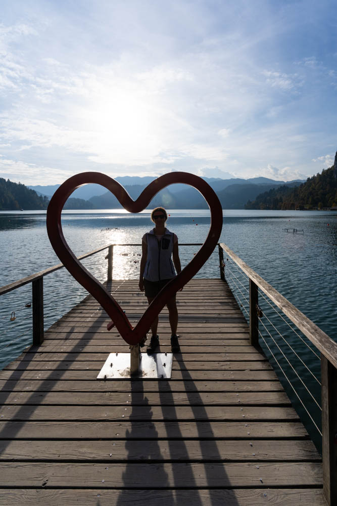 Auf einem Steg am Bleder See ist ein großes Herz aufgestellt. Melanie blickt durch das Herz in die Kamera. Dahinter sind der See Bled sowie die Berge zu sehen. Roadtrip Slowenien