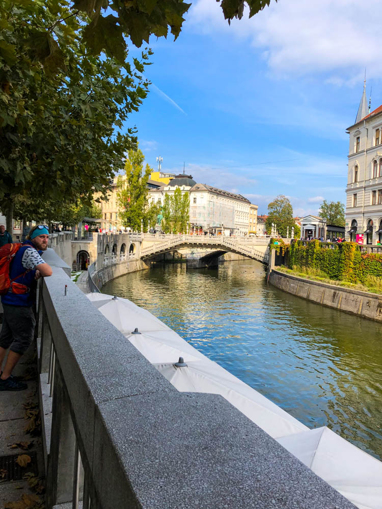 Julian lehnt an einer Mauer am Fluss. In der Mitte des Bildes sind die Drei Brücken zu sehen. Eine der Sehenswürdigkeiten in Slowenien - Roadtrip