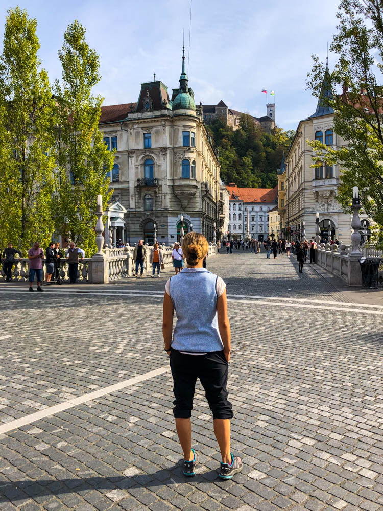 Melanie steht auf einem Platz in Ljubljana und blickt Richtung der Drei Brücken. Zudem ist die Burg der Stadt zu sehen. Roadtrip Slowenien