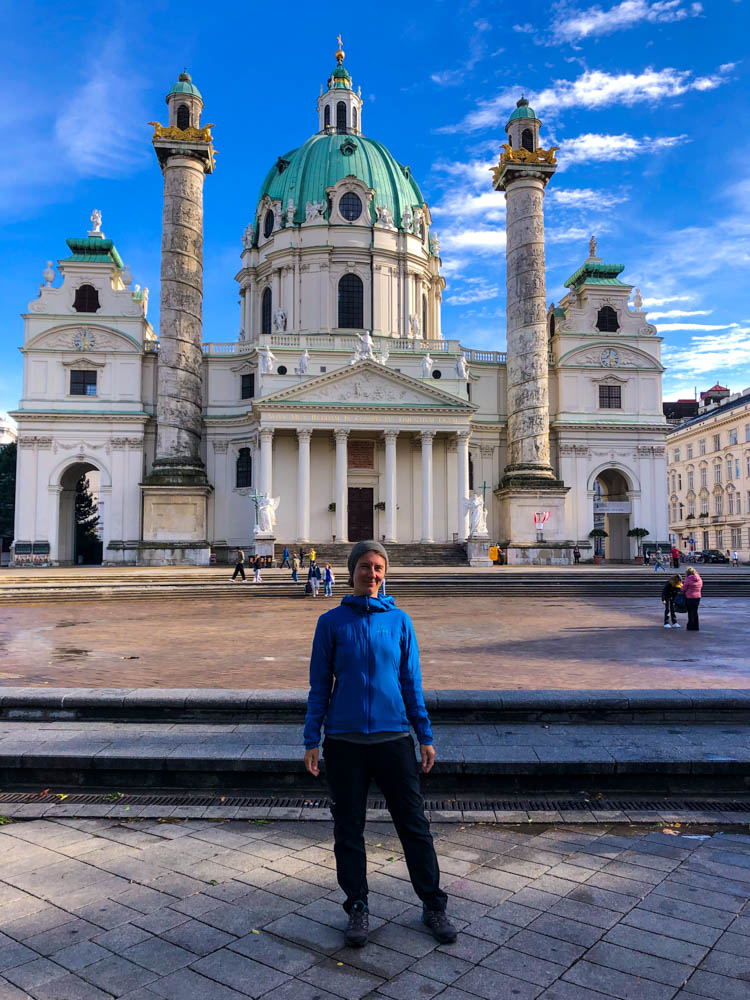 Melanie steht vor der Karlskirche in Wien und grinst in die Kamera. Der Himmel ist kräftig blau. Querfeldein durch Österreich in die Hauptstadt
