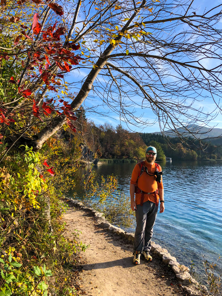 Julian steht neben einem See im Nationalpark Plitvicer Seen in Kroatien und grinst in die Kamera. Links vom Bild sind rot verfärbte Blätter an den Bäumen zu sehen. Der Himmel ist kräftig blau und nur leicht bewölkt.