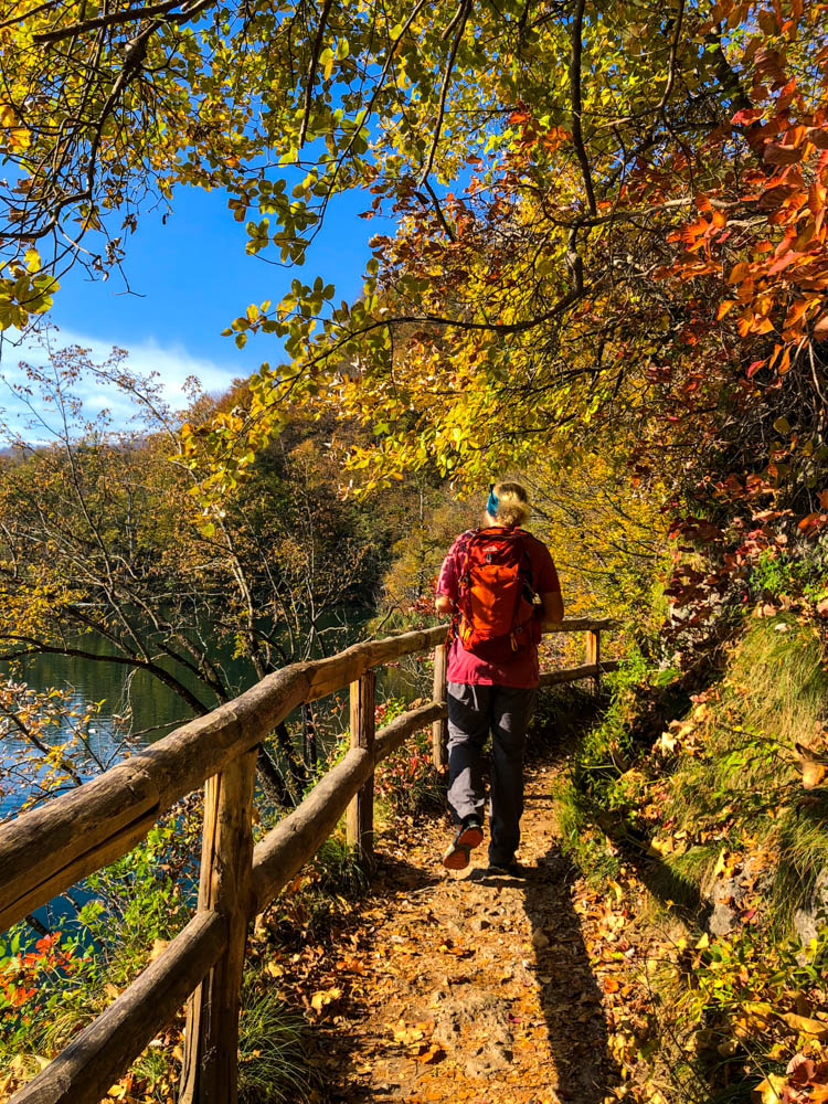 Julian wandert auf einem Weg neben einem See im Nationalpark Plitvicer Seen in Kroatien. Herbstliche Farben und kräftig blauer Himmel