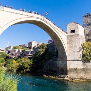 Titelbild für Text Bosnien Herzegowina - Ein Mann springt von der Stari Most in Mostar in den darunter fließenden Fluss. Der Himmel ist kräftig blau.