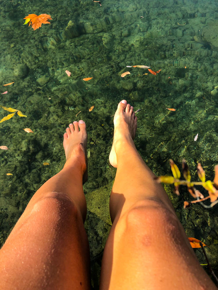 Melanie hängt ihre Füße in einen kalten Fluss, in welchem ein paar Blätter schwimmen