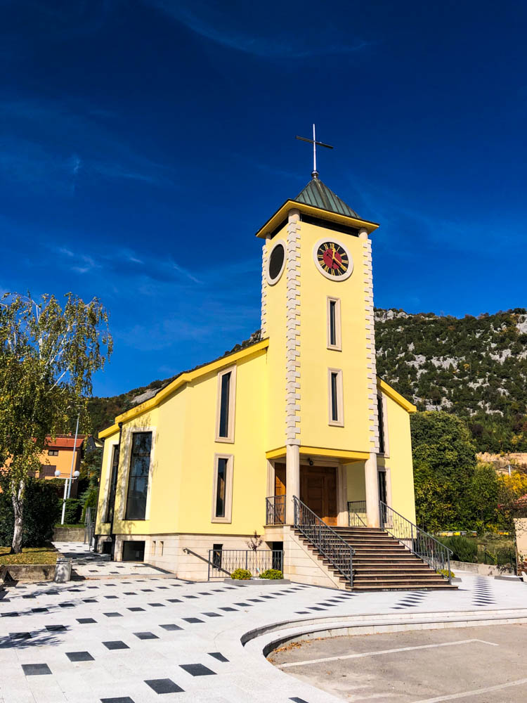 Kirche in einem Dorf in Bosnien und Herzegowina. Der Himmel ist kräftig blau und es ist etwas Natur rund um die Kirche zu sehen.