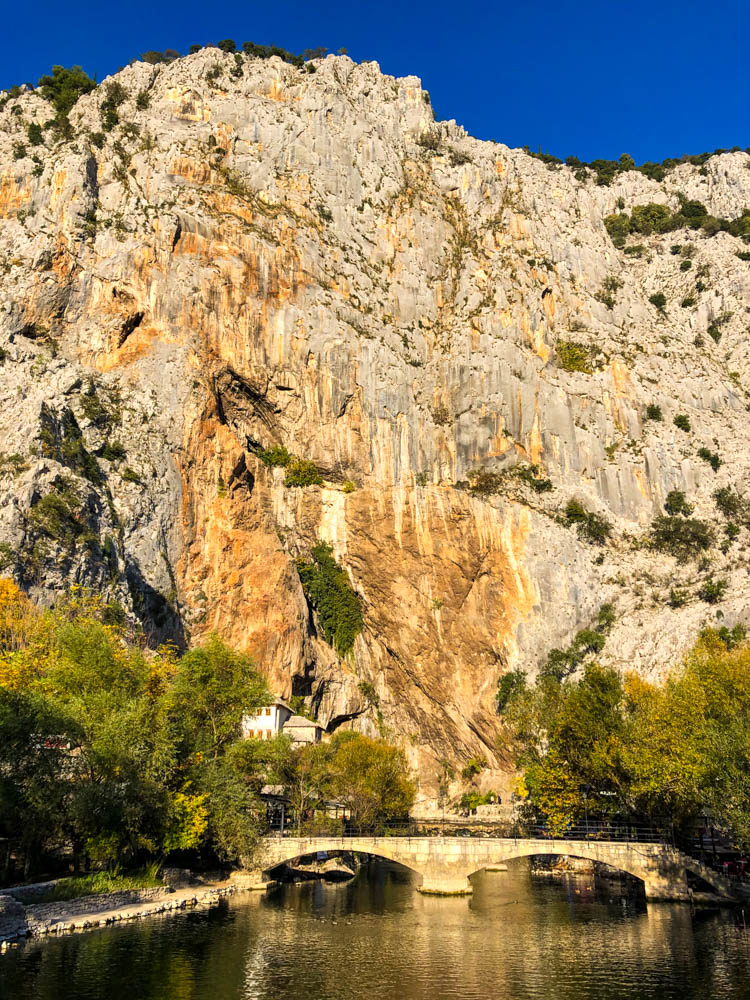 Steile Felswand in Blagaj in Bosnien und Herzegowina mit Derwischkloster. Es ist zudem eine Brücke über den Fluss zu sehen. Der Himmel ist kräftig blau