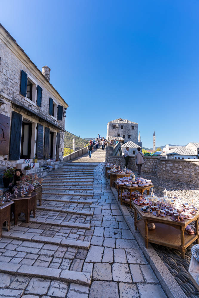 Kopfpflastersteine auf dem Weg zur Stari Most in Mostar, dem Wahrzeichen der Stadt. Rechts und links des Weges sind mehrere Marktstände zu sehen. Der Himmel ist kräftig blau.