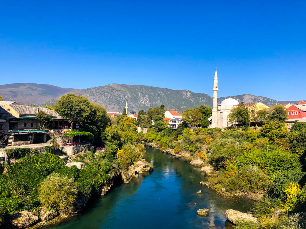 Blick von Stari Most auf Mostar in Bosnien und Herzegowina. Es ist im Zentrum des Bildes ein Fluss zu sehen, mehrere Moscheen sowie Häuser der Stadt. Der Himmel ist kräftig blau, am Horizont sind mehrere Hügel.