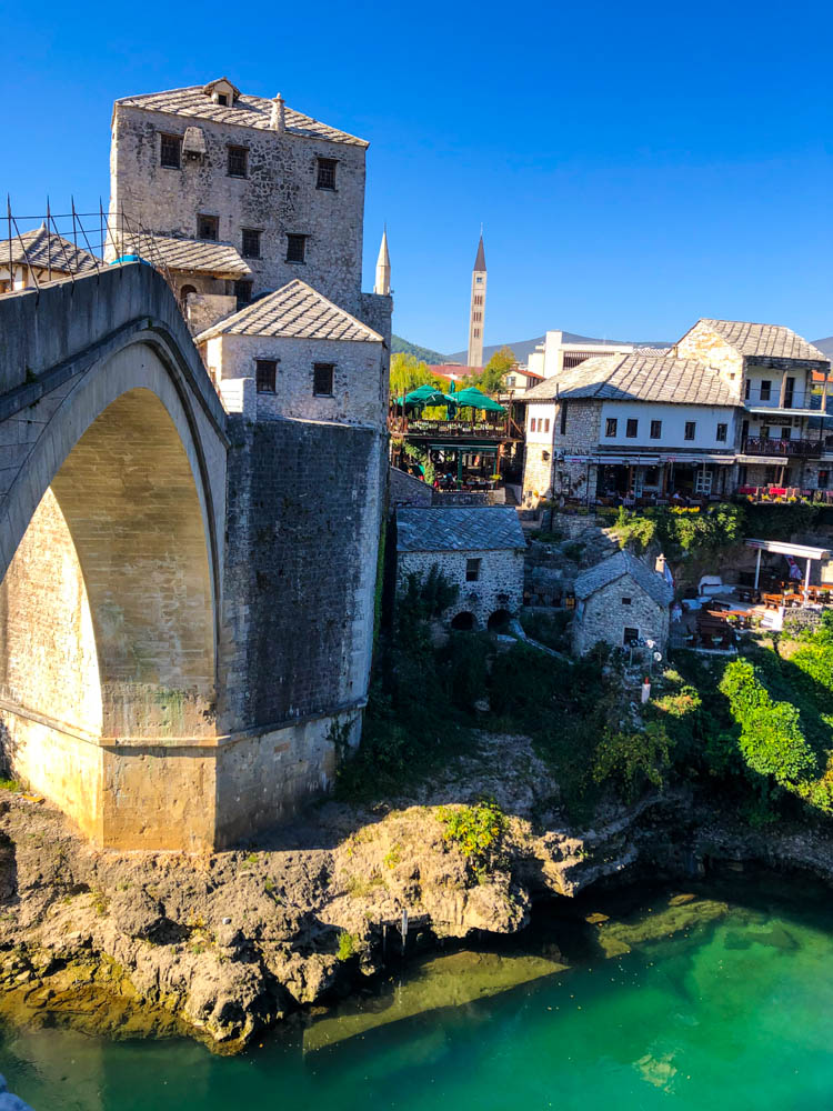 Stari Most in Mostar (Bosnien und Herzegowina). Hinter der Brücke ist eine Moschee zu sehen. Unter der Brücke fließt ein Fluss, der Himmel ist kräftig blau.