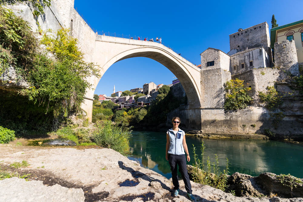 Melanie steht unterhalb der Stari Most in Mostar (Bosnien und Herzegowina), unter welcher ein Fluss durchfließt. Es ist die alte Stadtmauer zu sehen. Der Himmel ist kräftig blau.