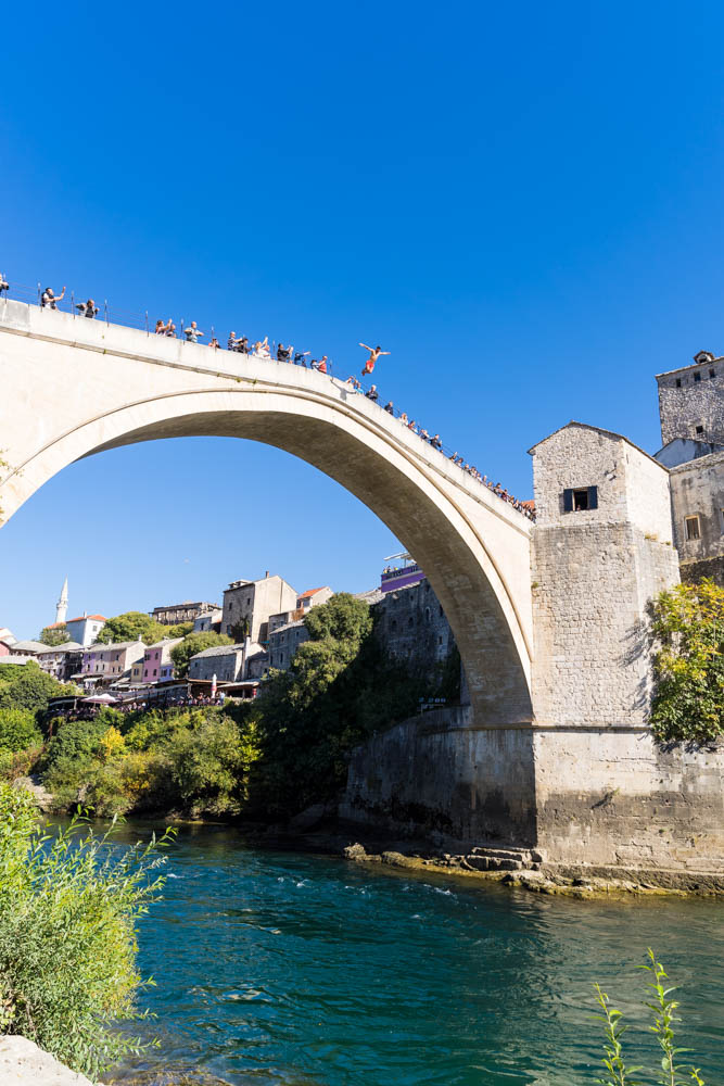 Ein Mann springt von der Stari Most in Mostar (Bosnien und Herzegowina) in den darunter fließenden Fluss. Der Himmel ist kräftig blau.