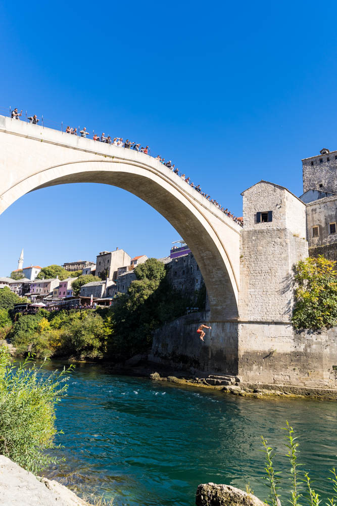 Ein Mann springt von der Stari Most in Mostar (Bosnien und Herzegowina) in den darunter fließenden Fluss. Der Himmel ist kräftig blau.