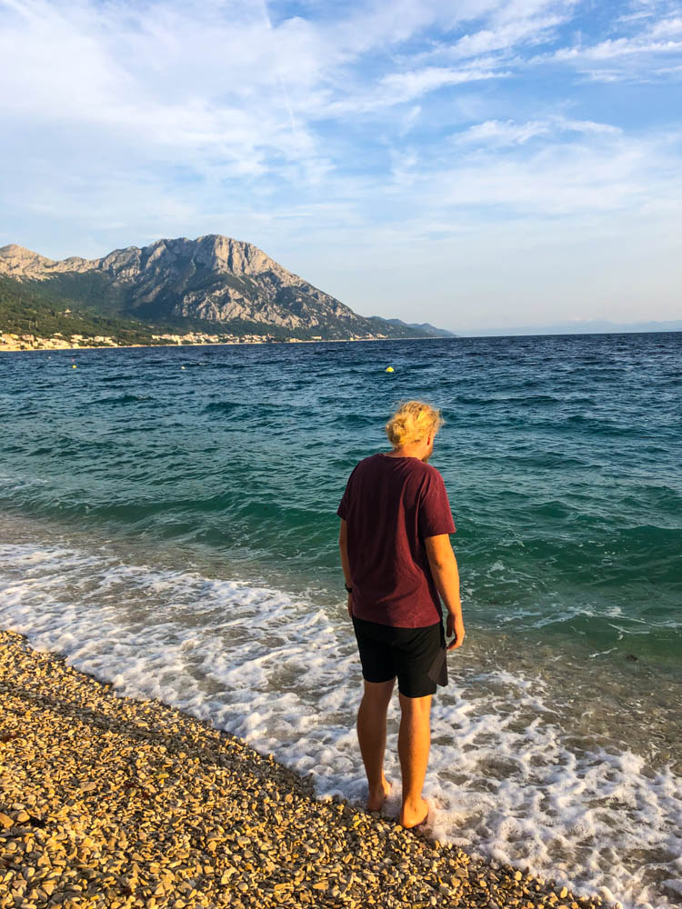 Julian steht mit seinen Füßen im Meer in Kroatien. Er wird von der Abendsonne angestrahlt und am Horizont sind ein paar Berge zu sehen.