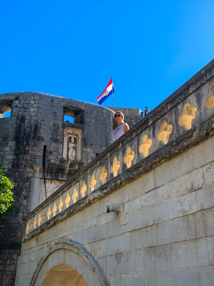 Melanie steht auf einer Mauer und blickt in die Kamera. Aufnahme von unten. Über Melanie ist die Stadtmauer zu sehen, eine kroatische Fahne sowie der kräftig blaue Himmel.