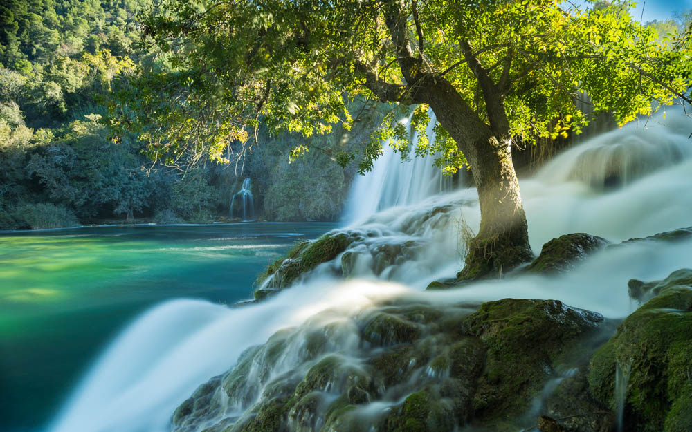 Langzeitbelichtung vom Wasserfall im Krka Nationalpark. Das Wasser ist verschwommen. Mitten im Wasserfall steht ein grüner Baum, welcher einen schönen Kontrast zum weiß und türkisen Wasser sowie dem blauen Himmel bietet.